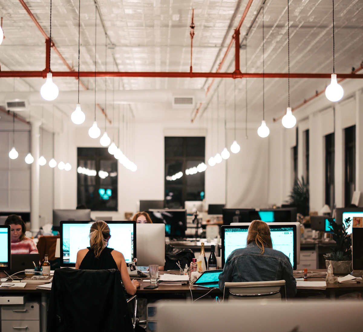 An open-plan office at night: people at their desks, each facing a large screen, under a row of pendant bulbs.